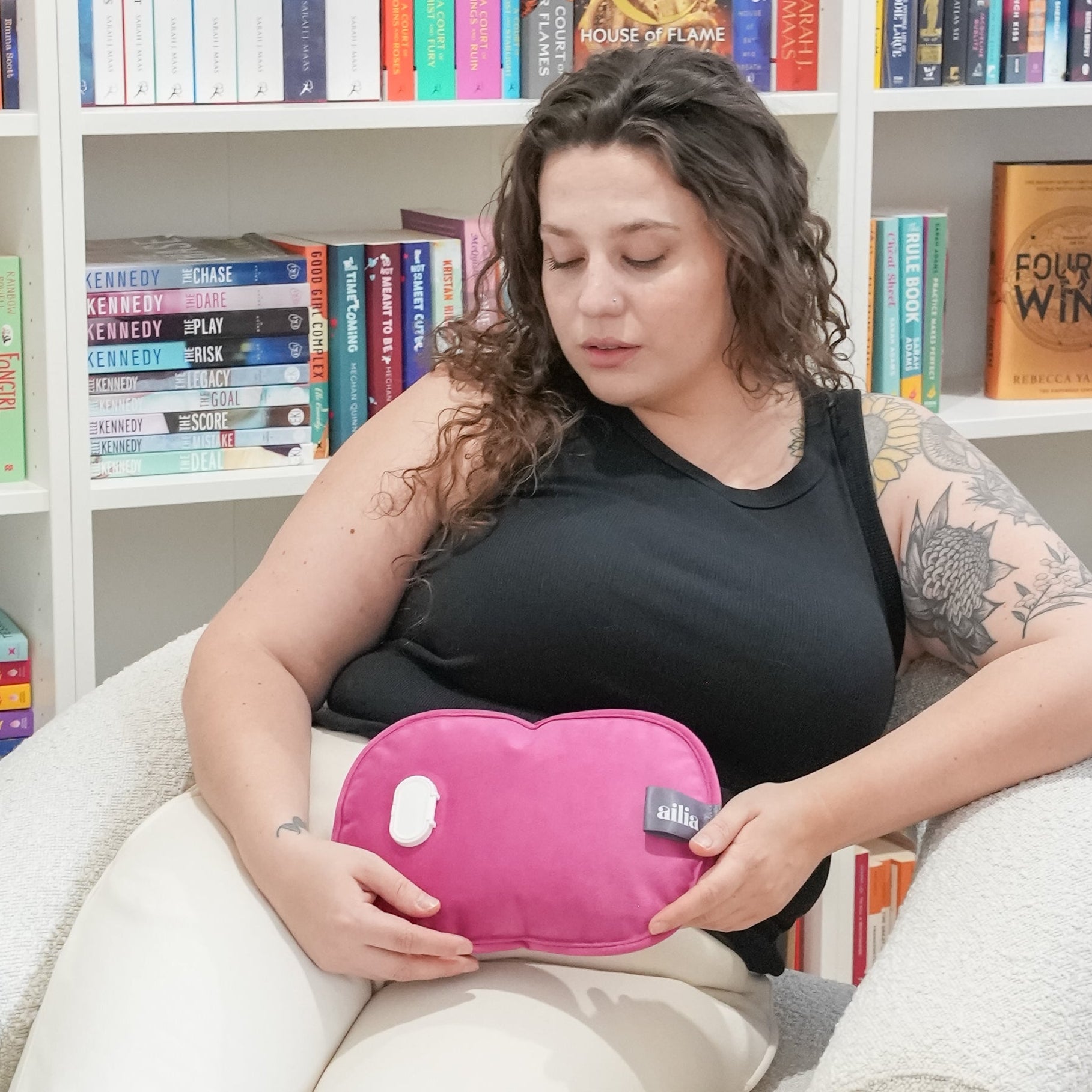 Woman sitting on a chair holding her pink ailia electric hot water bottle in front of a bookshelf.
