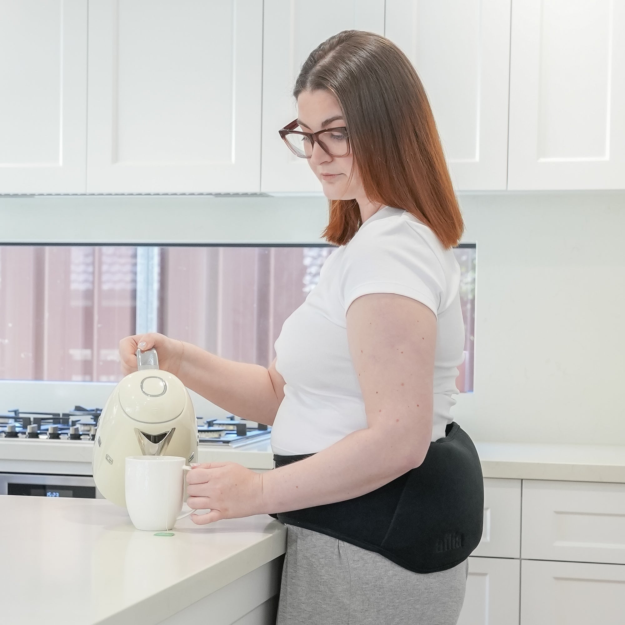 Woman making herself a cup of tea whilst wearing her Ailia Bottle Body Strap on her Lower Back.